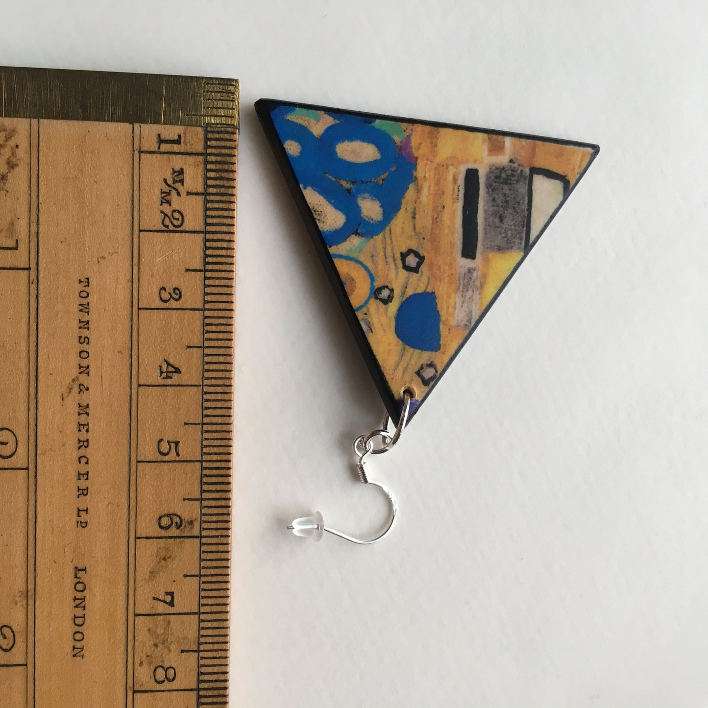 Klimt triangular earring nearby a rule that indicate the measurement of 4, 5 cm of his side.