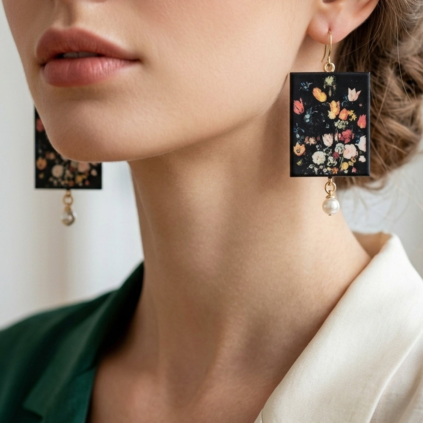 #A close-up photograph of a woman wearing large, rectangular handmade statement earrings. The earrings are black and feature a detailed, colorful Renaissance floral painting print inspired by Jan Brueghel. A small, elegant white pearl dangles from the bottom of each rectangular piece, attached with gold hooks. The woman is wearing a dark green top under a cream-white open collar shirt, with her hair styled in an elegant updo.