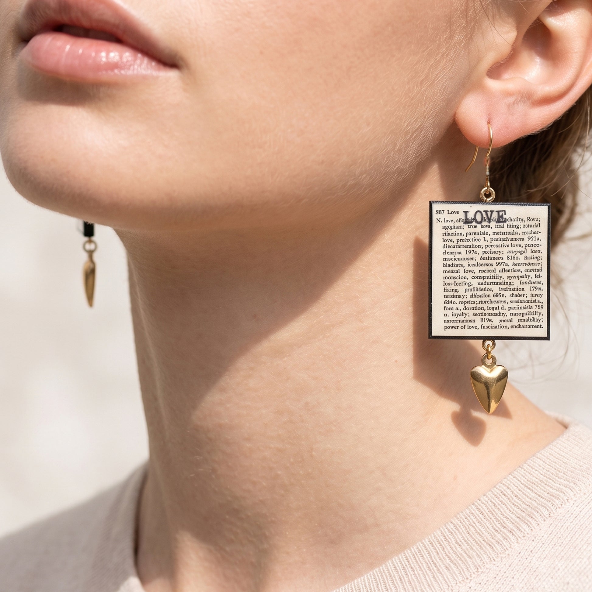 #A bright, close-up shot of a model from the lips down, wearing a pair of mismatched statement earrings. The earrings feature square pendants resembling dictionary pages with the word "LOVE" printed on them. The earring on the left side of the image has a smooth, solid gold heart charm dangling at the bottom, while the earring on the right features a black sacred heart charm with an ornate gold border. The model is wearing a high-necked white top against a clean, minimal background.
