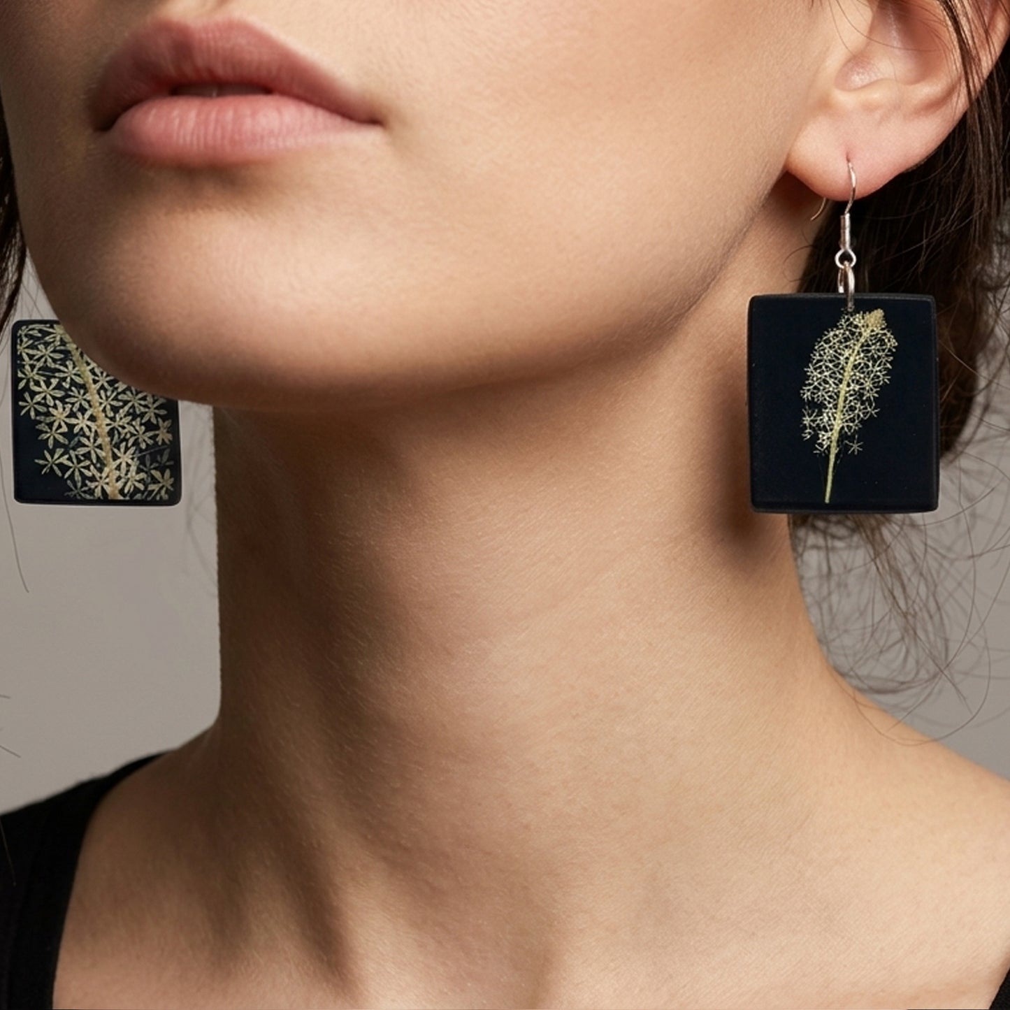 #Close-up of two square, matte black drop earrings with silver fishhook finders. The left earring displays a single stem of small white flowers, while the right one shows a denser cluster of blossoms, both styled like pressed botanical specimens.