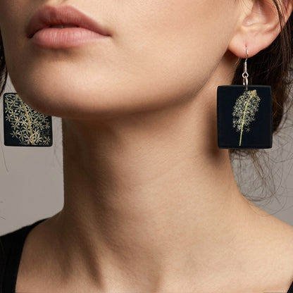 #Close-up of two square, matte black drop earrings with silver fishhook finders. The left earring displays a single stem of small white flowers, while the right one shows a denser cluster of blossoms, both styled like pressed botanical specimens.