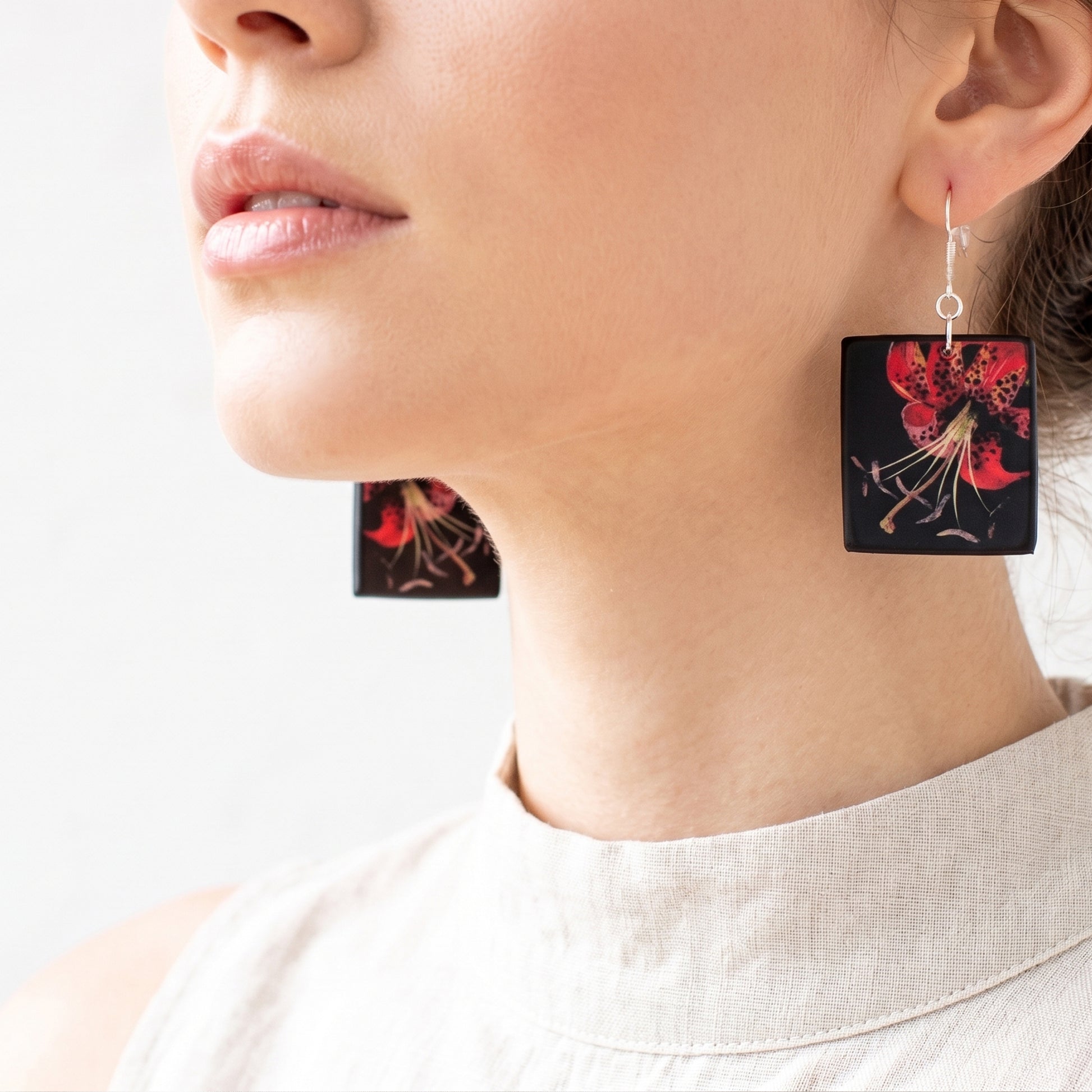 #Mary Delany art flowers square wooden earrings by Obljewellery. Close-up photo of a woman wearing black wooden earrings decorated with a vibrant red lily design, complete with custom silver engraved brand tags.