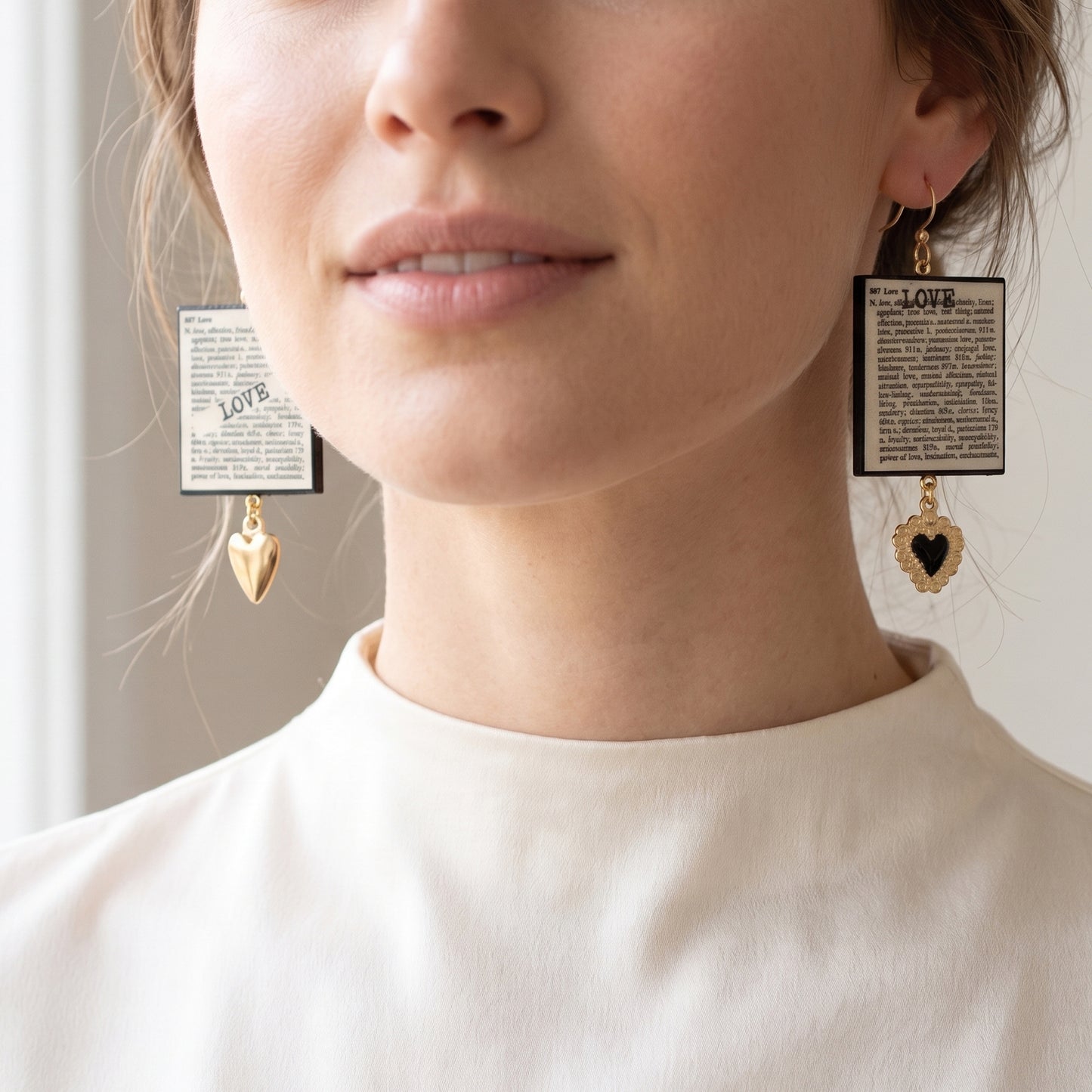 #A bright, close-up shot of a model from the lips down, wearing a pair of mismatched statement earrings. The earrings feature square pendants resembling dictionary pages with the word "LOVE" printed on them. The earring on the left side of the image has a smooth, solid gold heart charm dangling at the bottom, while the earring on the right features a black sacred heart charm with an ornate gold border. The model is wearing a high-neckeClose-up of a person wearing earrings with heart and rectangular designs.